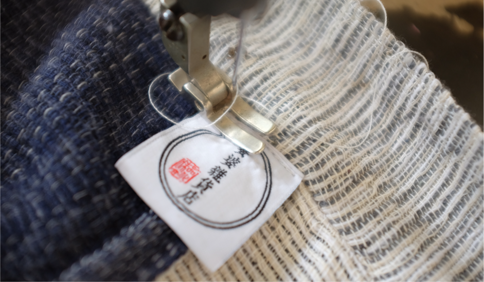 Extreme close-up of a sewing machine stitching the brand label onto the textured Hand-cranked Kimono at Grandmama Store in Hong Kong.