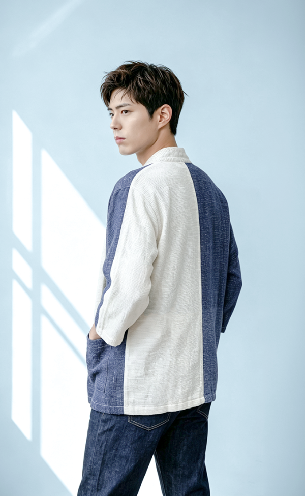 A young man with dark hair poses against a light blue wall with natural window light casting geometric shadows, viewed from the back-right profile. He wears a hand-cranked, color-blocked woven kimono jacket in Mt. Fuji blue and off-white with a visible collar label, paired with dark denim jeans at Grandmama Store in Hong Kong.