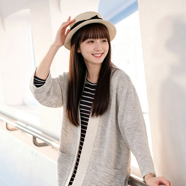 A smiling young woman with long brown hair and bangs wearing a straw fedora hat, standing outdoors next to a railing while wearing a hand-cranked Kimono cardigan in the Aurora Snow colorway—featuring a heavily textured cream and light gray slub-knit handwoven cotton fabric, wide cuffs, and a solid cream trim—styled open over a navy and white striped top at Grandmama Store in Hong Kong.