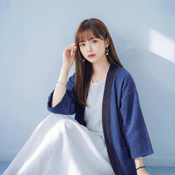 A young woman with straight brown hair and bangs sitting down while wearing a hand-cranked Kimono cardigan in Moonlight color—featuring a heavily textured medium blue handwoven body, wide three-quarter length sleeves, and a contrasting charcoal gray collar trim—styled open over a simple white dress against a soft, light blue wall backdrop at Grandmama Store in Hong Kong.