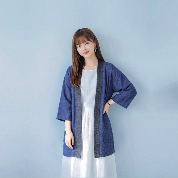 A young woman with long straight brown hair and bangs smiling at the camera while posing with one hand on her hip, wearing an open-front hand-cranked Kimono cardigan in Moonlight color—featuring a heavily textured medium blue handwoven cotton body, three-quarter length wide sleeves, and a contrasting charcoal gray collar trim—styled over a white dress against a clean, light blue wall backdrop at Grandmama Store in Hong Kong.