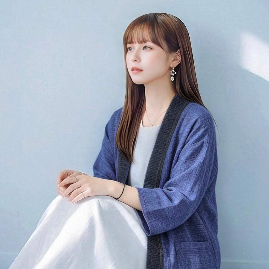 A young woman with long straight brown hair and bangs looking off to the side while sitting down, wearing an open-front hand-cranked Kimono cardigan in Moonlight color—featuring a heavily textured medium blue handwoven cotton body, three-quarter length wide sleeves, and a contrasting charcoal gray collar trim—styled over a white dress against a clean, light blue wall backdrop at Grandmama Store in Hong Kong.