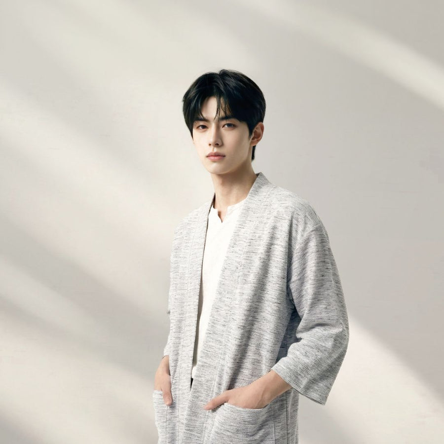 A young man with black hair posing with his hands in his pockets, wearing an open-front hand-cranked Kimono cardigan in the Rockstream colorway—featuring a highly textured heather gray and white speckled slub-knit handwoven cotton fabric, wide sleeves, and front patch pockets—styled over a plain white inner shirt against a minimalist off-white backdrop with soft shadows at Grandmama Store in Hong Kong.
