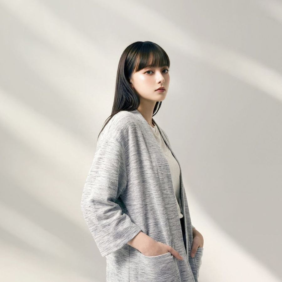 A young woman with straight black hair and bangs posing in profile with her hands in her pockets, wearing an open-front hand-cranked Kimono cardigan in the Rockstream colorway—featuring a highly textured heather gray and white speckled slub-knit handwoven cotton fabric, wide sleeves, and front patch pockets—styled over a simple light cream top against a minimalist off-white backdrop with soft shadows at Grandmama Store in Hong Kong.
