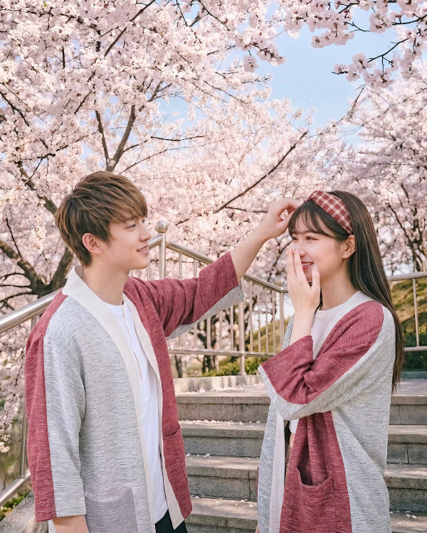 A young couple wearing matching hand-cranked Kimono cardigans in Tokyo Tower color—featuring a dusty-red body, light gray sleeves, and cream-white collar trim—sharing a sweet moment on outdoor stone steps framed by blooming pink cherry blossom trees at Grandmama Store in Hong Kong.