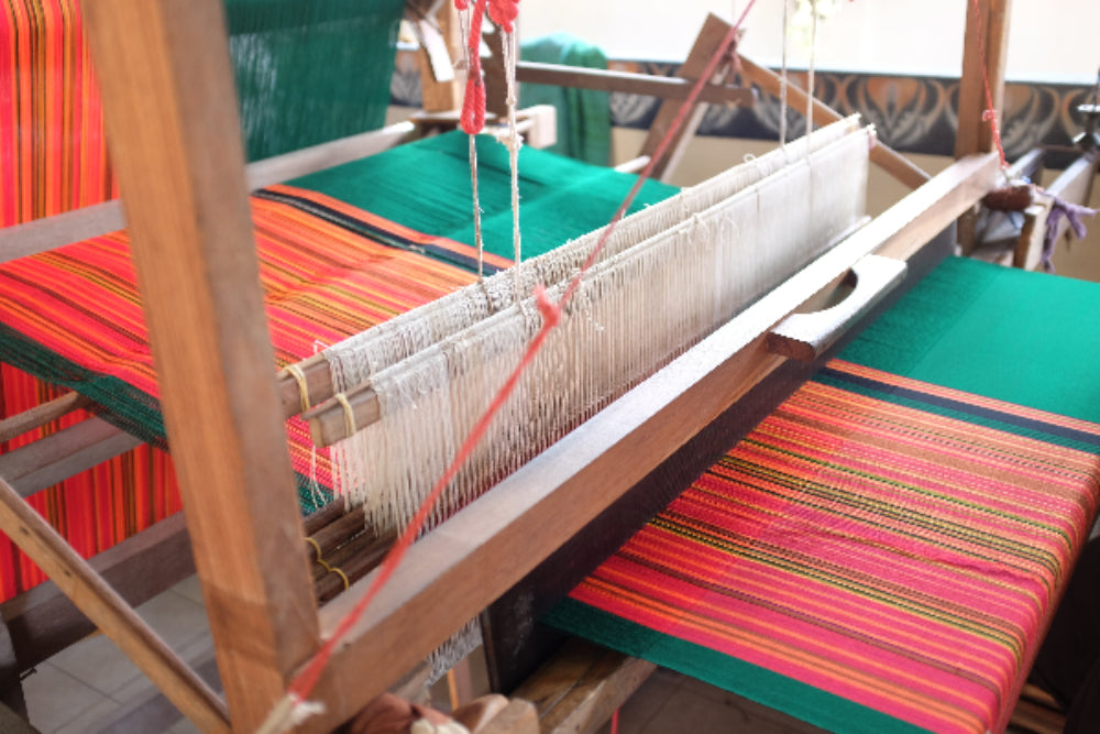 A close-up shot of a traditional wooden handloom in action, showing vibrant, freshly woven textile with colorful pink, orange, and yellow stripes contrasted against sections of solid forest green fabric, with the white cotton heddles and warp threads intricately strung through the loom frame at Grandmama Store in Hong Kong.