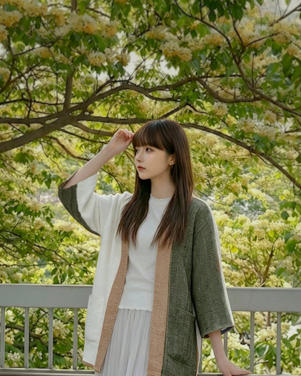 A young woman with straight brown hair and bangs wearing a hand-cranked Kimono cardigan in Yakushima color—featuring a vertical split design with textured forest green on one side, cream-white on the other, and a warm tan woven collar—posing outdoors in front of a lush tree blooming with pale yellow flowers at Grandmama Store in Hong Kong.